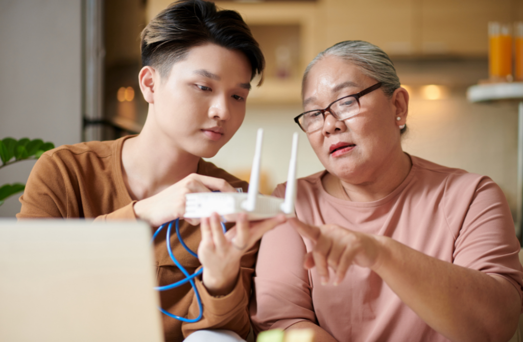 A young man and an older woman are sitting together at a table, examining a Wi-Fi router. The man holds the device, while the woman points at it. They appear focused, indoors with a laptop and kitchen items visible in the background.