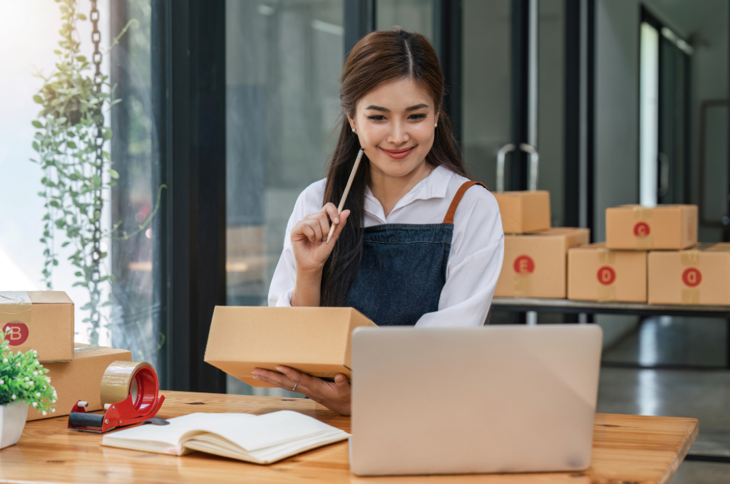 Woman sitting at a table holding a cardboard box, smiling at a laptop. Packing materials, tape, and boxes are on the table. She appears to be working in a small business or online store setting.