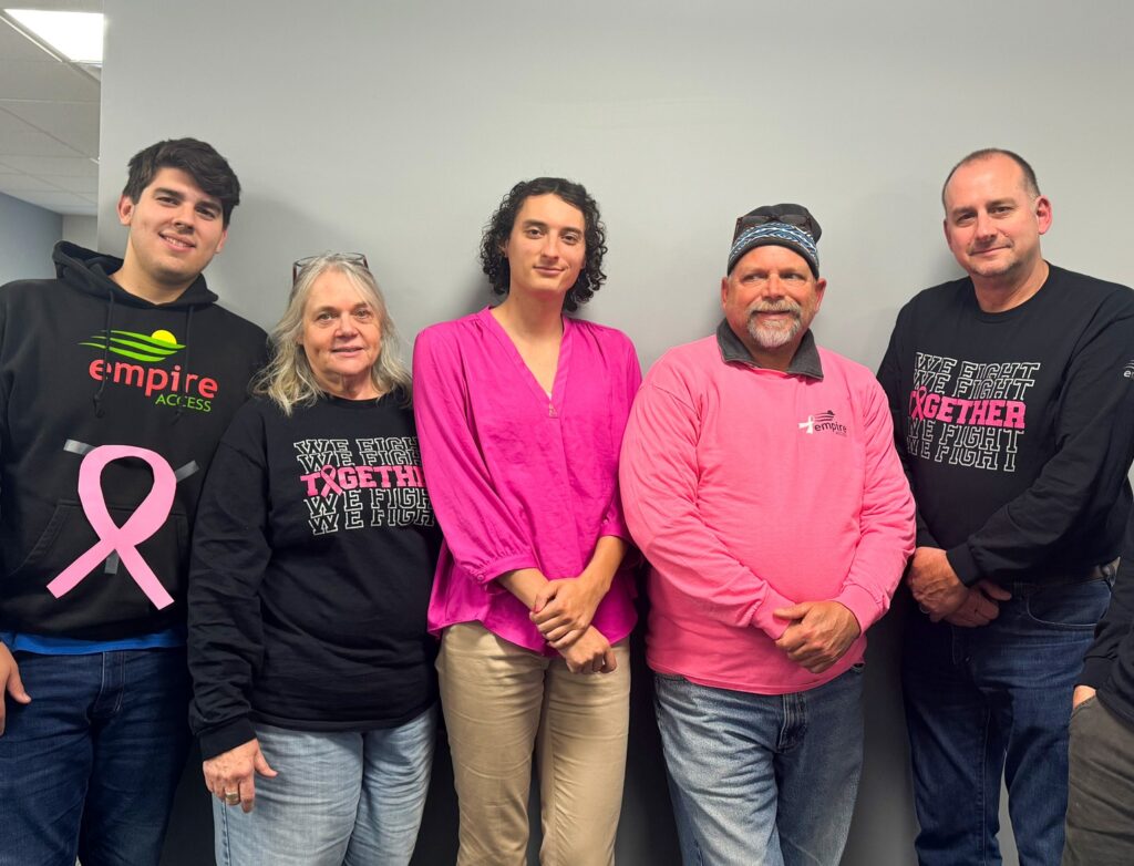 A group of seven people stand together indoors, some wearing black shirts with a pink breast cancer awareness ribbon and the phrase "We Fight Together, We Fight." One person is wearing a bright pink shirt. They are posing against a gray wall.