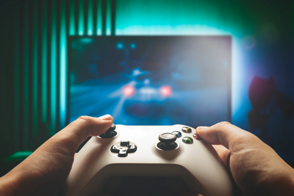 Hands holding a white video game controller in front of a blurry, illuminated screen displaying a racing game. The room has a dark ambiance with a green and blue glow.