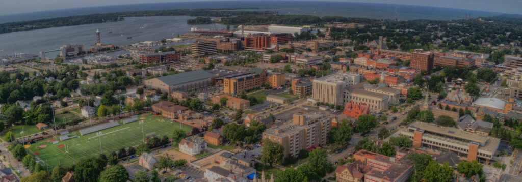 Aerial view of a coastal city with Fiber Internet in Erie, showcasing modern and historical buildings, a sports field in the foreground, tree-lined streets, and a bustling harbor with boats. The backdrop reveals a vast body of water and distant land on the horizon.