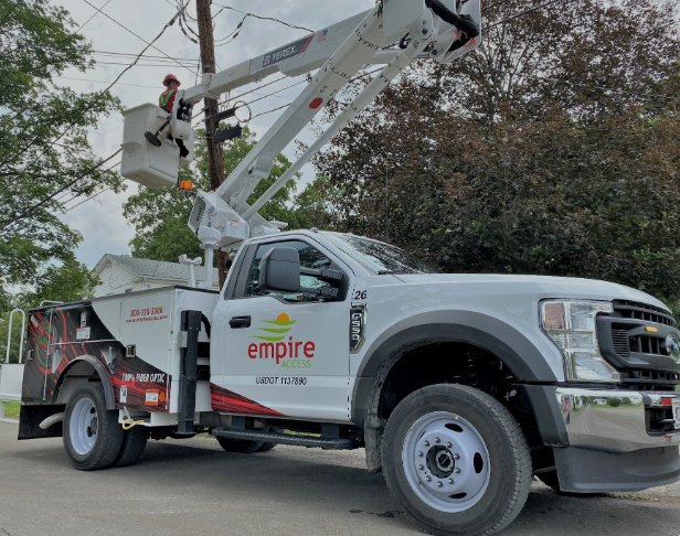 A white utility truck with "Empire Access" branding is parked on a street, showcasing their commitment to connectivity. A worker, emblematic of the values found on their About Us page, is elevated in a safety-equipped bucket. Trees and power lines stretch across the background.