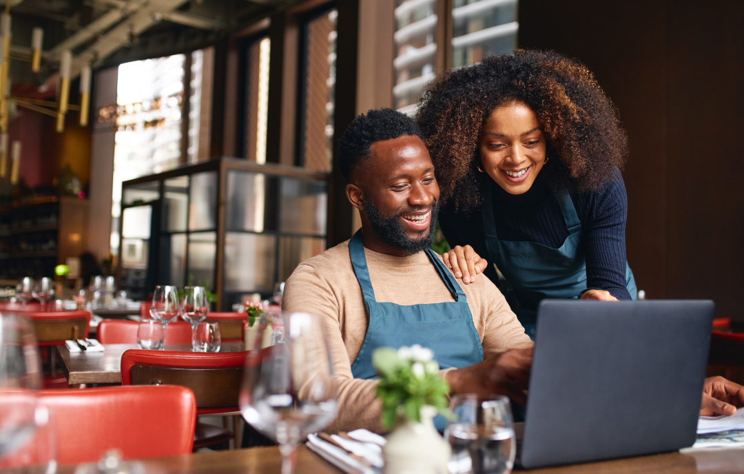 A man and woman, both wearing aprons, stand behind a laptop in a cozy restaurant setting. They are smiling and appear engaged with the screen. The table is set with glasses and a small plant centerpiece.