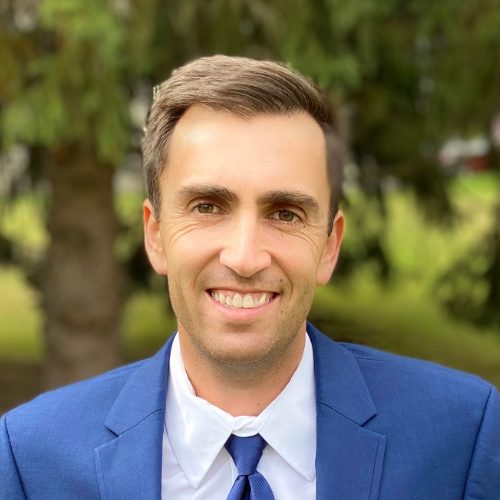 A person wearing a blue suit and tie smiles confidently, standing outdoors with blurred greenery in the background, embodying the professionalism and warmth you'd expect from Empire Access.