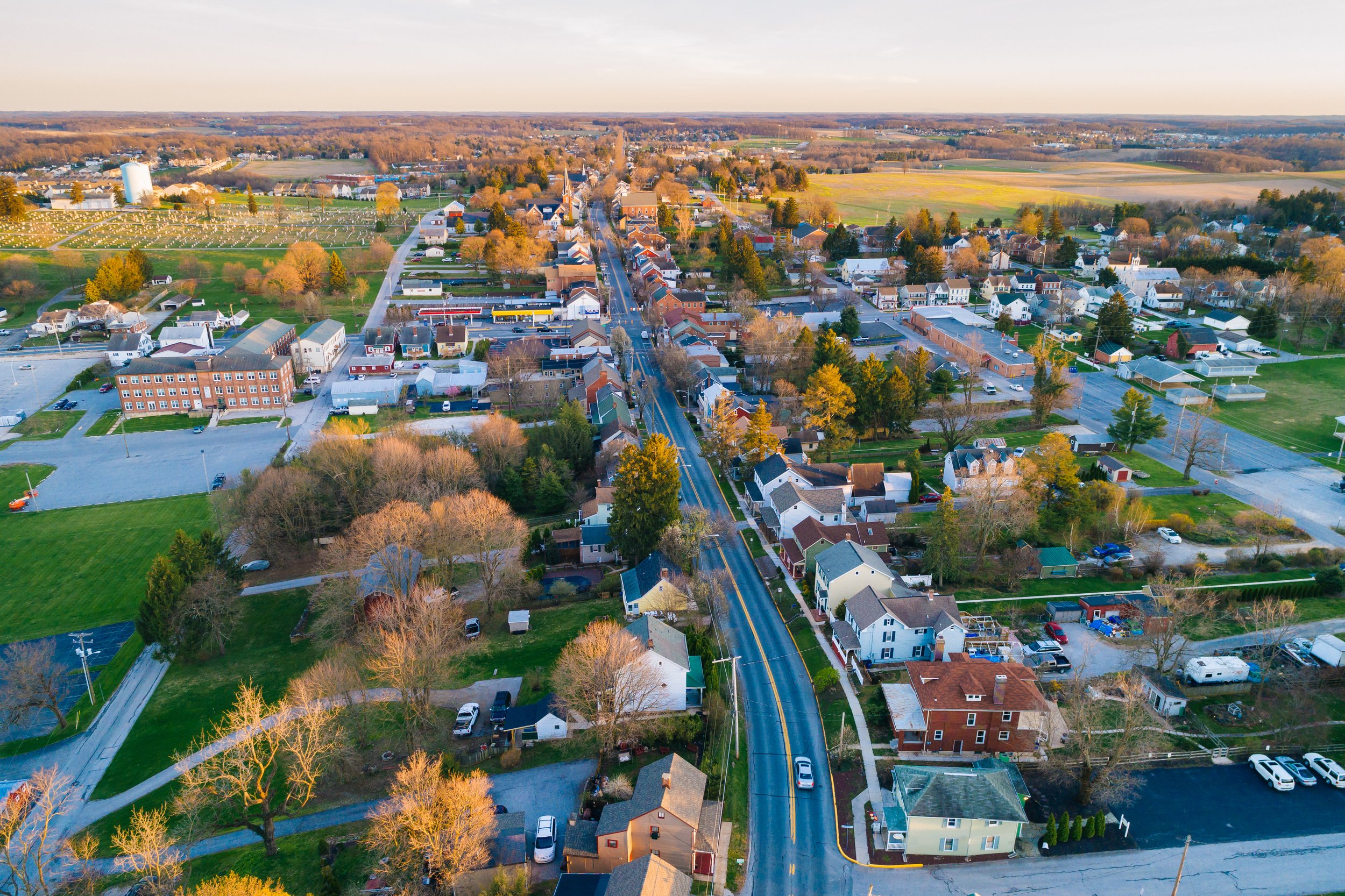 Aerial view of a small town at sunset with a main road running through the center. Houses, trees, and commercial buildings are visible, surrounded by vast fields and open spaces under a clear sky.