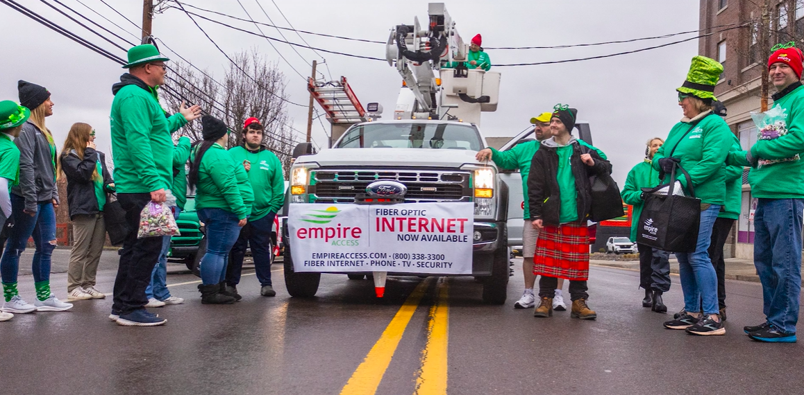 People in green attire stand near a white utility truck with an "Empire Access" banner promoting fiber-optic internet. The scene is festive, suggesting a parade or public event. The backdrop includes a building and utility equipment.